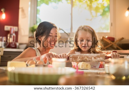 A mother and her four years old daughter are cooking in a luminous kitchen. They are working on a pasty with a rolling pin on a wooden table full of ingredients and a green baking pan. Shot with flare