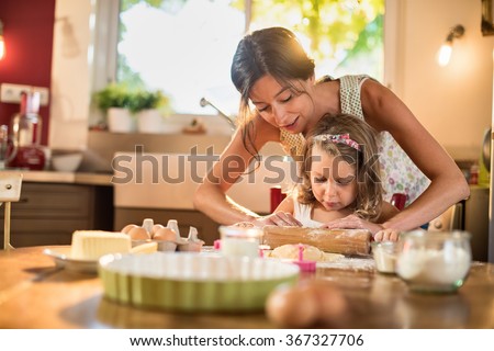 A mother and her 4 years old daughter are cooking in a luminous kitchen. They are working on a pastry with a rolling pin on a wooden table full of ingredients and a green baking pan. Shot with flare