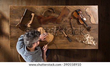 Top view. Craftsman luthier working on the creation of a violin. He sits at a wooden table in his workshop, various tools and instrument are placed on the workbench