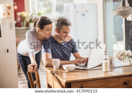 Nice thirty year couple using a laptop while having breakfast in the kitchen, they are wearing casual clothes and th man has gray hair