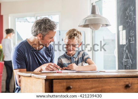 Young boy doing his school homework with his father, the boy is writing in his book, at the blurred background the mother works in the kitchen