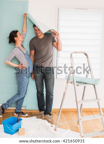 Funny couple, a pregnant woman and his husband wallpapering the bedroom walls of their future baby, a piece of wallpaper falling on dad\'s head