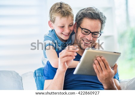 At home, a father and his young son having fun by gaming on a tablet, dad sits on a white couch and the boy looks at the screen over the shoulder of his father and giving him advices to win
