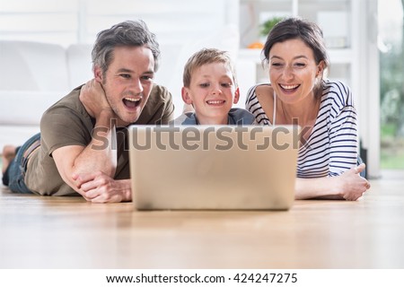 At home, cheerful family sharing a funny video on a laptop. They are lying on the wooden floor of the living room.
