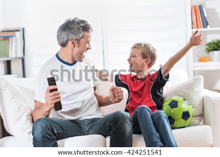 At home, father and his son watching TV with a ball at their side, they raise their arms in victory, daddy has the remote in hand