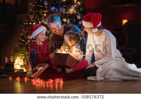 Christmas night. Near the christmas tree a lovely family opening their gifts. They enjoy the warm Christmas atmosphere in their living room, mom and kids wearing a hat of Santa Claus.