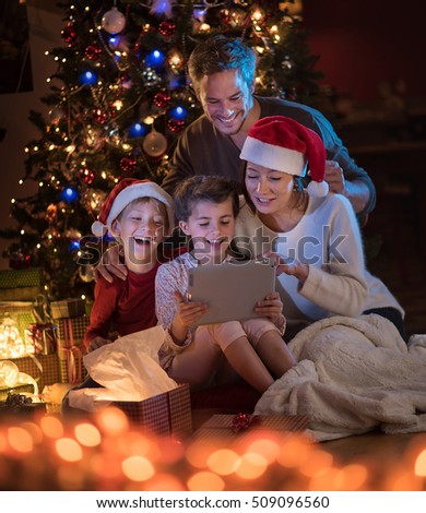 Christmas night. Near the christmas tree a lovely family opening their gifts. they are very happy to find a digital tablet inside. Mom and kids wearing a hat of Santa Claus.