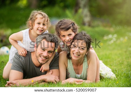 Cheerful family in a park, parents and their two lovely daughter are lying on the grass while they are looking at camera. Shot with flare