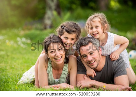 Cheerful family in a park, Dad, mum and heir two lovely daughter are lying on the grass while they are looking at camera. Shot with flare