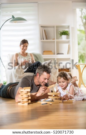 Cheerful family at home, Daddy and his young daughter lying on the wooden floor are playing with a wooden game.  At background mom is sitting in an armchair while using a digital tablet