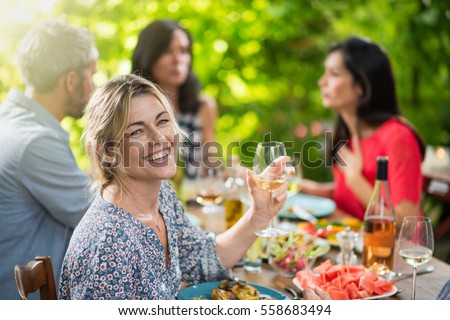 Looking at camera, portrait of a beautiful middle aged blond woman sharing a meal friends on a terrace table in summer, she has a glass of wine at hand. Shot with flare