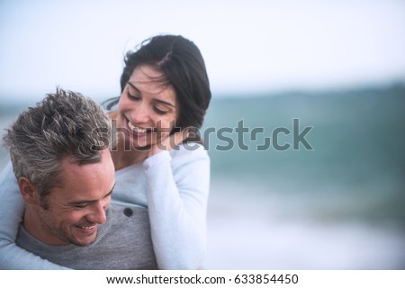 A beautiful couple in their forties walks on the beach, the man wears the woman on his back, they wear sweaters and jeans