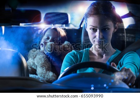 Front view. Portrait of a beautiful woman driving her car at night in the rain. In the back her little girl fell asleep her teddy in the arms