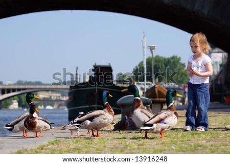 Little beauty girl feedind wild ducks at landing stage or river embankment at sunny day. We see ship not in fokus at background. Prague, Vltava.