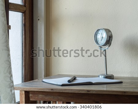 Workplace with notebook, pen, clock on a wooden table. And foreground is curtains blowing in the wind