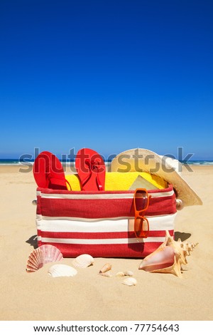 Beach bag with book, seashells, sunglasses, hat and towel in a deserted beach at a summer day with deep blue sky