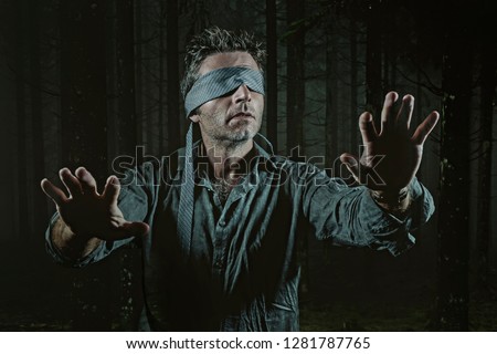 young confused and scared man blindfolded with necktie playing internet trend dangerous viral challenge with eyes blind lost in dark forest background guided by intuition