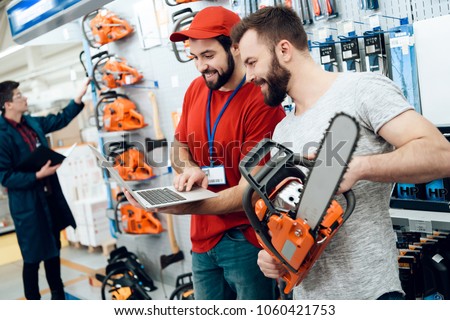 Salesman in red shirt and baseball cap is showing bearded client new chainsaw explaining details on laptop in power tools store.