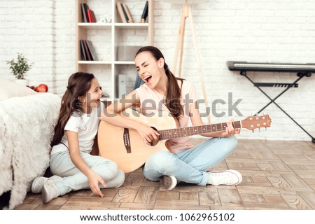 Mom and her daughter are sitting on the floor at home and playing the guitar. They sing to the guitar. Behind them is a bookshelf, a synthesizer and a white wall.