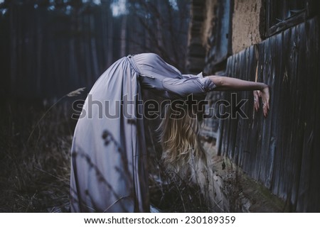 Blonde young model in a lavender maxi dress bends back in a creepy unnatural way in the dark forest