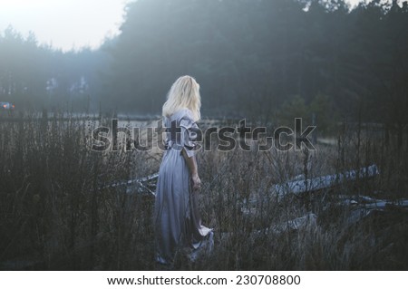 Blonde young model in a lavender maxi dress in the dark creepy forest