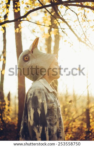 man half took off his rubber bird mask, standing sideways in the autumn sunset forest thinking dramatically