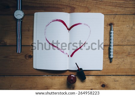 Paper notebook with a heart drawn on it with a red nail polish, watch, pen, red nail polish on the wooden background