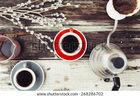 Fresh morning black coffee in a red cup with a red saucer, a kettle  and a dripper with a used coffee filter on the wooden old background and some pussy willow