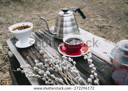 Fresh morning coffee in a red cup on the old wooden table with a kettle, coffee dripper and some pussy willow outdoor picnic