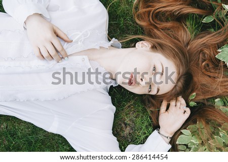 Beautiful redhead woman lying on the fresh green grass wearing a white vintage shirt