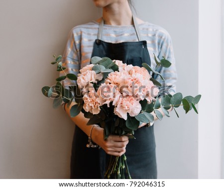 Young florist woman holding freshly made minimalistic blossoming flower bouquet of pastel pink carnations and eucalyptus on the grey wall background.