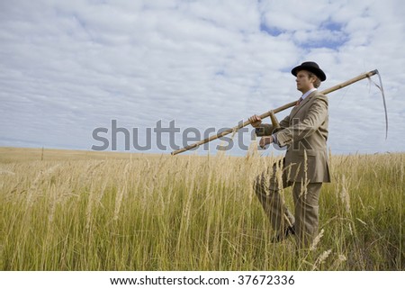 man in suit and hat with scythe go in field with golden grass