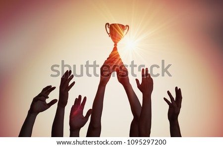 Winning team is holding trophy in hands. Silhouettes of many hands in sunset.