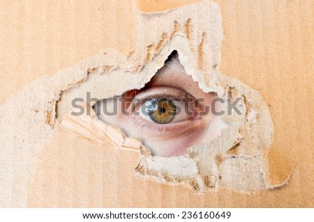 Hidden eye spying through torn hole in cardboard paper