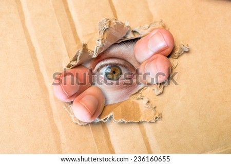 Hidden eye spying through torn hole in cardboard paper