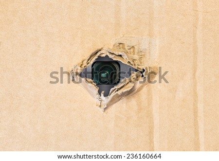 Hidden camera lens spying through torn hole in cardboard paper