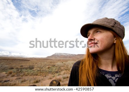 Wide angle close up of pretty, young red headed woman wearing hat with snow covered mountain and hill in background. Early winter.
