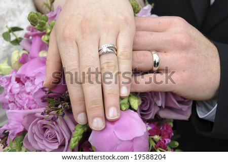 A bride and groom show off their wedding rings by placing their left hands on the bride\'s bouquet