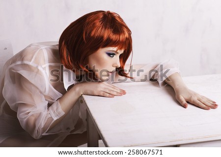 Young woman with red hair sitting near the table and thinking.  Bob hair cut. Red hair. White table. Sitting and posing. Fashion model