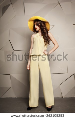 Beautiful young woman with curly long brown hair standing in yellow hat and jumpsuit.  Fashion model. Stylish young people. Young beautiful woman with long hair. Beautiful hairstyle. Yellow hat.