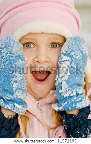 surprised young girl outside in the snow wearing a pink hat and blue snow covered mittens.