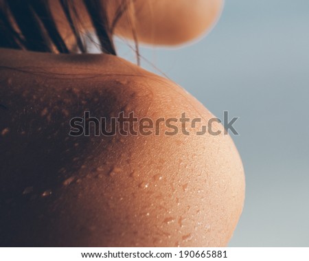 Close up texture of the wet shoulder of a young woman showing skin detail and the way the water condenses and beads on the surface of the skin against a sunny blue summer sky