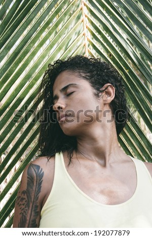 Beautiful girl standing against a tropical palm frond with her head turned to the side with closed eyes and a serene expression