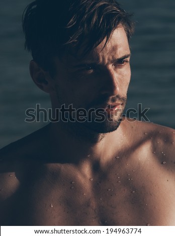 Close-up portrait of a sexy handsome Caucasian young man, bearded and tanned, posing shirtless while looking aside, in a hot sunny day at the seaside