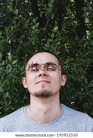 Young man with a goatee beard and a peaceful serene expression closing his eyes in bliss as he tilts his head back with a smile of pleasure against a leafy tree with copyspace