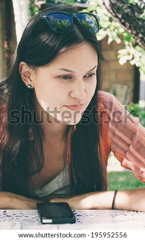 Attractive young woman sitting at a table outdoors dreaming as she waits for a call on her mobile phone looking thoughtfully off to the right of the frame
