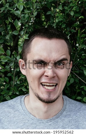 Angry young man with a goatee beard baring his teeth and snarling at the camera with a raised eyebrow and frowning expression, head and shoulders against greenery outdoors