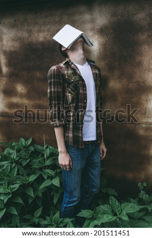 Man standing with his head tilted back and a book covering his face in lush green shrubs against an old wall