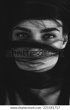 Portrait of a mysterious woman with her long brown hair wrapped in strands across her face looking at the camera with seductive luminous eyes, monochrome head and shoulders image