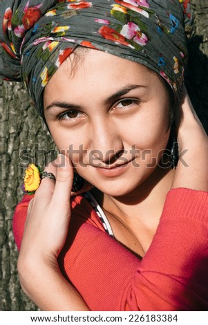 Close up Smiling Pretty Young Woman with Fashion Floral Head Cover Leaning on Tree While Crossing Arms. Looking at Camera.